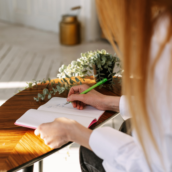 Image: a woman sitting at a desk with the focus on her hands. One hand is holding a pen, the other is holding a journal open. The image includes flowers and decor that feels boho and cozy. Symbolizing the idea of journaling to answer the question Am I An Alcoholic