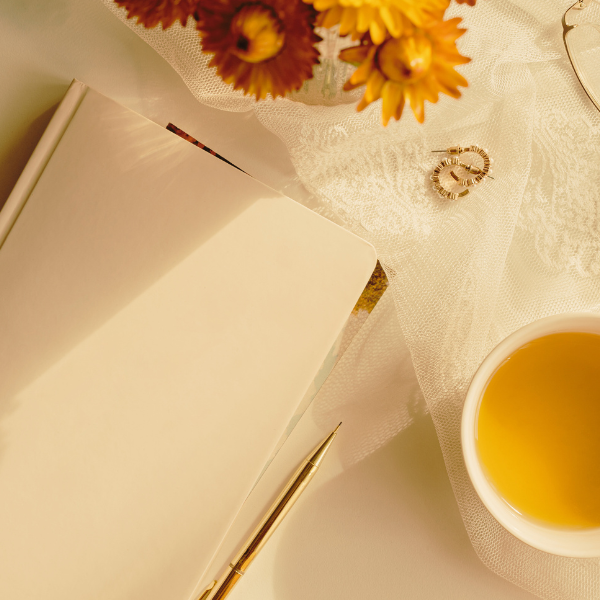 image: A white journal with a gold pen laying on a table. Surrounding the journa are some red and yellow flowers, a white mug of yellow tea, and a pair of golden earrings. 