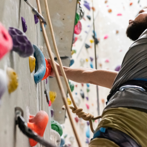 Image: A close up of a man wearing a gray shirt and yellow shorts, wearing a rock climbing harness. Colorful grips can be seen all around. Image illustrates one of many "sober winter adventures" that the author is describing. 