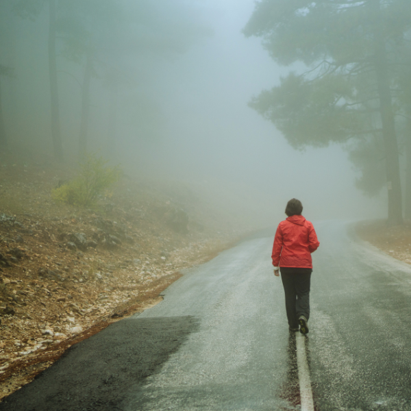 Image: Woman wearing a red jacket and black pants, walking alone down a road, surrounded by mist and trees. Only mist can be seen in the distance. Image represents the feelings of isolation and shame that come with alcoholism.